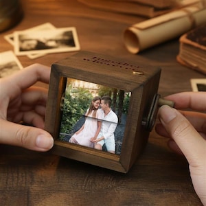 May include: A wooden music box with a photo of a couple inside. The box is square with a crank on the side and the words "together wherever" engraved on top. The box is held by a person's hands.