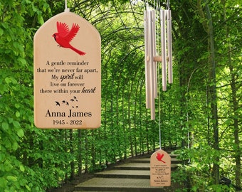 May include: A wooden wind chime with a red cardinal and the text "A gentle reminder that we're never far apart, My spirit will live on forever there within your heart Anna James 1945 - 2022" hanging from a tree branch.