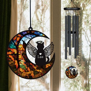 May include: A stained glass wind chime featuring a black cat with angel wings and a halo, sitting inside a crescent moon. The moon is decorated with autumn leaves in shades of orange, yellow, red, and green. The cat's name, "Leo", is written below the heart on its chest.