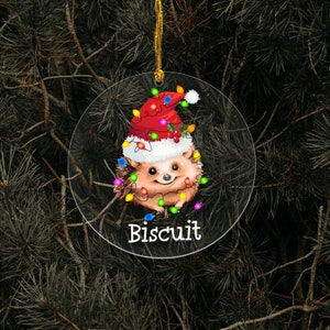 May include: A round, clear Christmas ornament with a cartoon hedgehog wearing a Santa hat and surrounded by colorful Christmas lights. The text "Biscuit" is printed below the hedgehog.