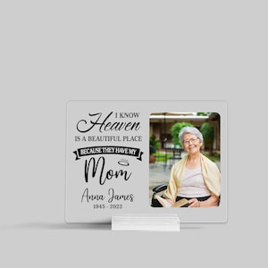 May include: A white memorial plaque with a black photo frame and a black inscription that reads "I know Heaven is a beautiful place because they have my Mom Anna James 1945 - 2022". The photo shows an older woman smiling.
