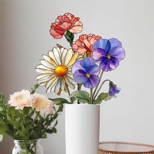 May include: A vibrant stained glass floral arrangement in a white vase. The bouquet features a daisy, carnations, and pansies in shades of red, orange, yellow, and purple. The vase is cylindrical.