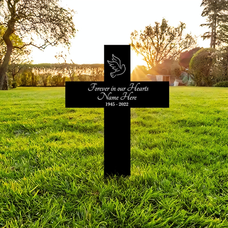 K&ouml;nnte beinhalten: Schwarzes Holzkreuz mit einer wei&szlig;en Taube und dem Text "Forever in our Hearts Name Here 1945 - 2022" auf einem gr&uuml;nen Rasen.