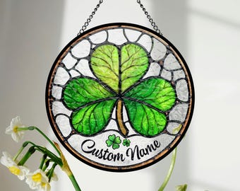 May include: Round stained glass window with a shamrock design. The shamrock is green with detailed leaf veins, set against a white background with a black border. The text "Custom Name" is written in cursive. A chain is attached for hanging.