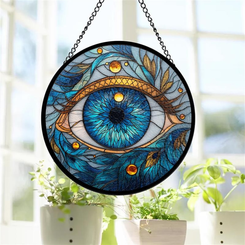 May include: A round stained glass window featuring a detailed eye design. The eye is primarily blue with gold and teal accents, set against a light background. The window is framed in black and suspended by a chain.