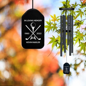 May include: Black wind chime with a black tag that reads "In Loving Memory 1960 - 2022 Kevin Marler" with a golf ball and crossed golf clubs graphic.