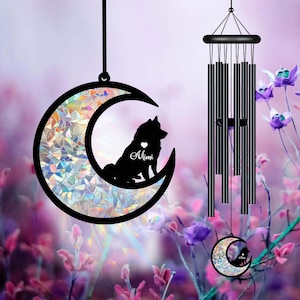 May include: A black metal wind chime with a crescent moon design. The moon has a holographic iridescent finish and a silhouette of a dog inside. The dog is sitting and has the name "Mimi" written below it. The wind chime is hanging from a white ceiling.