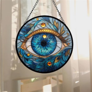 May include: Round stained glass window art featuring a detailed blue eye design. The eye is surrounded by blue and gold leaf patterns, with a black border and chain for hanging. The artwork is a decorative piece.