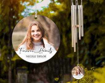Puede incluir: Un adorno redondo blanco para memorial con una foto de una joven y el texto "Forever in our Hearts Nicole Taylor". El adorno está colgado de una cuerda y está rodeado de follaje verde.
