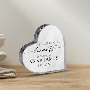 May include: A clear glass heart-shaped memorial with the text "Forever in our hearts" and "In Memory of ANNA JAMES 1945-2022" printed on it.