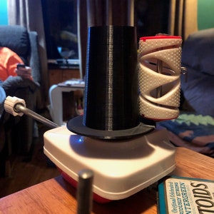 May include: A white yarn winder with a black plastic spool holder and a white plastic yarn guide. The winder is sitting on a wooden table with a tin of Altoids mints.