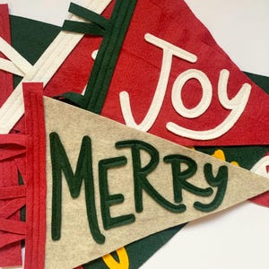 Puede incluir: Primer plano de varias pancartas de fieltro en rojo, verde y beige. Las pancartas presentan las palabras "Joy" y "Merry" en blanco y verde. Las pancartas tienen adornos decorativos y están diseñadas para la decoración navideña.