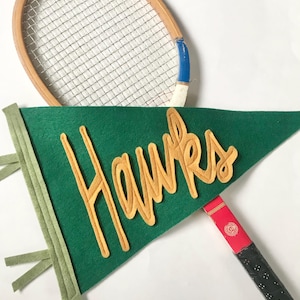 May include: A green felt pennant with the word "Hawks" in gold felt letters. A wooden tennis racket with blue and white trim is partially visible behind the pennant.