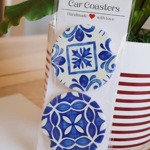 May include: Two car coasters with blue and white floral designs are packaged together. The coasters are round with a small notch and are made of ceramic. The packaging reads "Car Coasters Handmade with love."