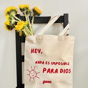 May include: A white canvas tote bag with red text that reads "Hey. Nada es imposible para Dios" and a red drawing of a smiling sun. The text "gaec" is also written in red below the sun.