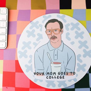 May include: A round mousepad with a blue background and a white pattern of X's. The mousepad features a cartoon illustration of a man with glasses and a mustache holding a cup. The text "Your Mom Goes To College" is printed in black at the bottom of the mousepad.