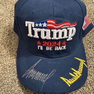 May include: A navy blue baseball cap with the word "Trump" in white, and the text "2024 I'll Be Back" in red. The cap features an American flag design and a signature.