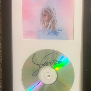 May include: Framed album art and CD. The album art shows a woman with blonde hair against a pink and blue backdrop, with the word "Lover" in cursive. Below is a CD with a signature.