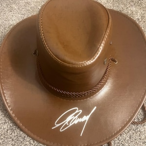 Puede incluir: Sombrero de vaquero marrón con ala ancha y firma blanca. El sombrero tiene una cinta de cuerda marrón y una correa para la barbilla. El sombrero está hecho de un material liso.