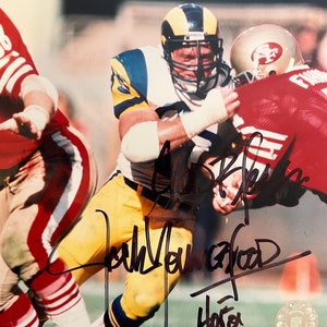 Puede incluir: Una fotografía en blanco y negro de un jugador de fútbol americano que lleva un uniforme amarillo y azul con un casco azul. El jugador está siendo tackleado por un jugador que lleva un uniforme rojo y blanco. La foto está firmada por el jugador con tinta negra. La firma dice "John  Youngblood" y "Horton".