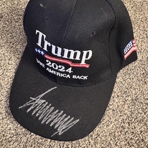 May include: A black baseball cap featuring white embroidered text that reads Trump 2024 Take America Back with a small American flag graphic. The cap includes a silver-colored embroidered signature on the brim and a small American flag on the side.