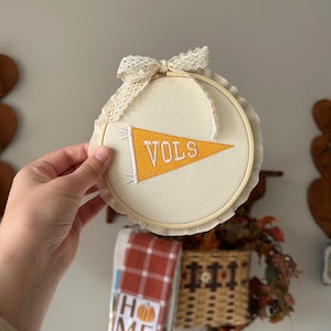 May include: A round, cream-colored embroidery hoop with a banner that reads "VOLS" in orange and white. The hoop is adorned with a lace bow and trim. The background includes a basket and a towel that says "HOME".