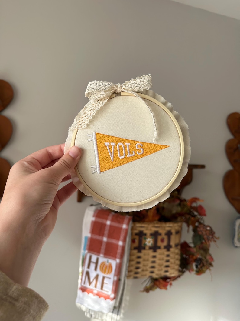 May include: A round, cream-colored embroidery hoop with a banner that reads "VOLS" in orange and white. The hoop is adorned with a lace bow and trim. The background includes a basket and a towel that says "HOME".