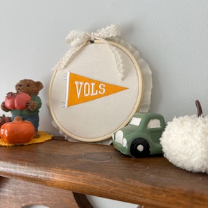 May include: A decorative wooden shelf displays a collection of fall-themed items. A cream-colored embroidery hoop with an orange pennant that reads "VOLS" is the focal point. Other items include a green toy car, a white pom-pom, and a small bear holding a pumpkin.