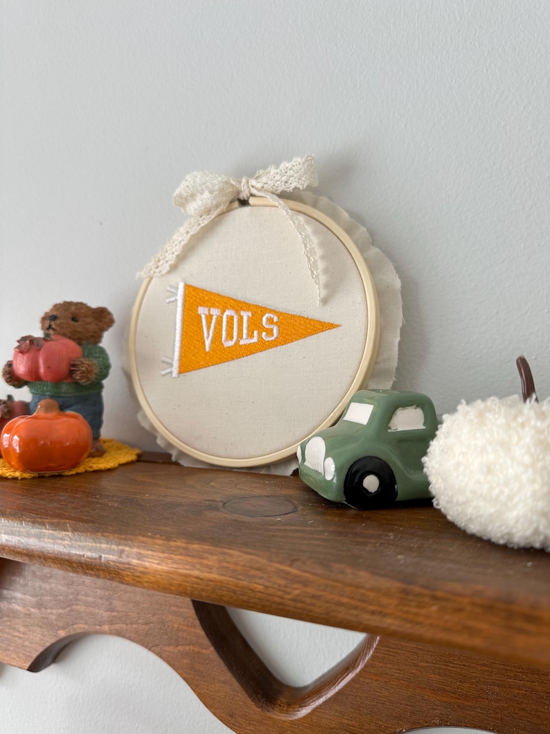 May include: A decorative wooden shelf displays a collection of fall-themed items. A cream-colored embroidery hoop with an orange pennant that reads "VOLS" is the focal point. Other items include a green toy car, a white pom-pom, and a small bear holding a pumpkin.