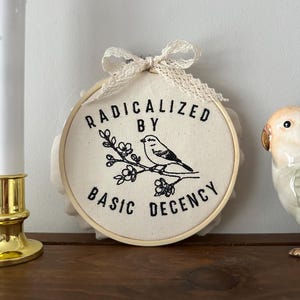 May include: A round, cream-colored embroidery hoop with the text "RADICALIZED BY BASIC DECENCY" in black. A black and white bird and floral design is embroidered below the text. The hoop is topped with a cream-colored lace bow.