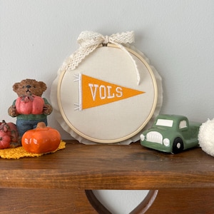 May include: A cream-colored banner with the word "VOLS" in orange, displayed within a wooden embroidery hoop. The banner is adorned with a lace bow. Decorative items include a green truck, a bear figurine, and orange pumpkins.