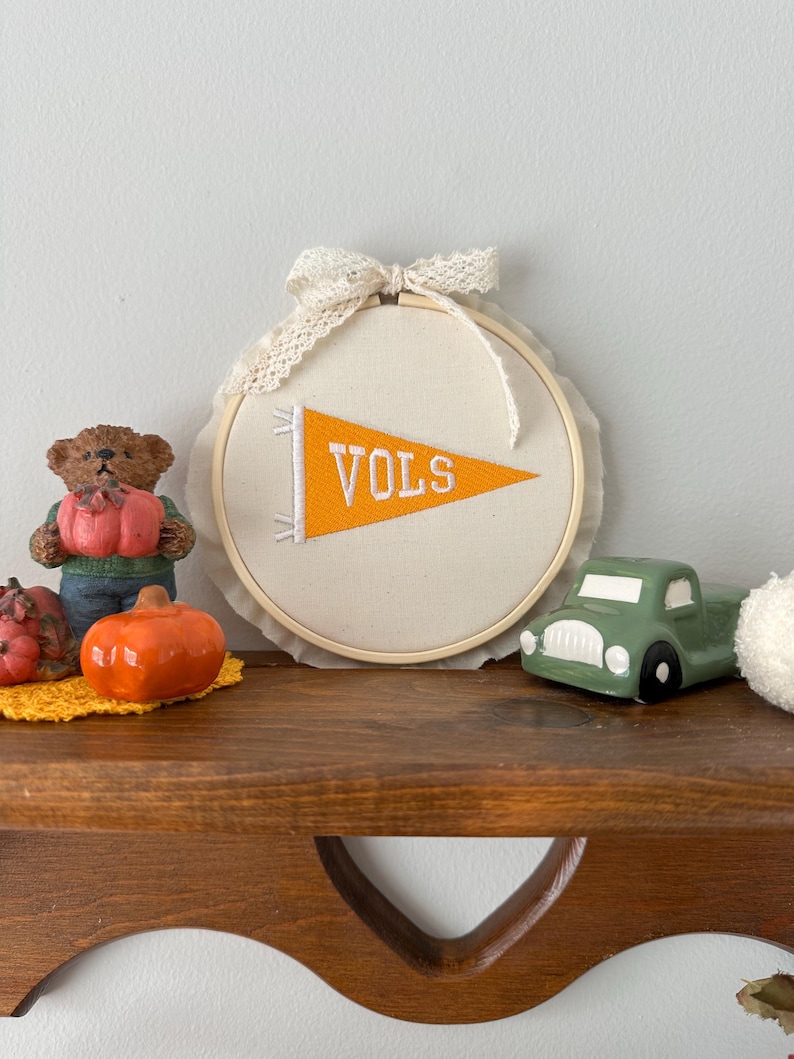 May include: A cream-colored banner with the word "VOLS" in orange, displayed within a wooden embroidery hoop. The banner is adorned with a lace bow. Decorative items include a green truck, a bear figurine, and orange pumpkins.