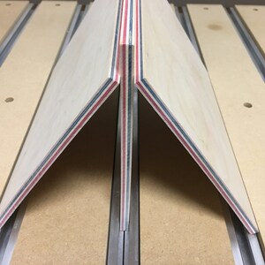 May include: Two pieces of wood with red, blue, and white stripes are joined together at a 90-degree angle. The wood is clamped to a table with a metal track.