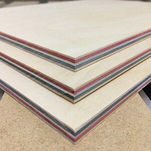 May include: Three pieces of plywood stacked on top of each other. The plywood has a light wood color with red, blue, and black stripes along the edges.