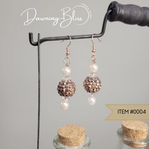 May include: Rose gold earrings with a sparkly brown ball and a white pearl bead. The earrings are hanging from a black metal stand. The text "Dawning Bliss" is visible in the top left corner of the image. The text "ITEM #0004" is visible in the bottom right corner of the image.