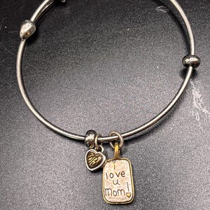 K&ouml;nnte beinhalten: Silberfarbenes Armreif-Armband mit Herz- und rechteckigen Anh&auml;ngern. Der rechteckige Anh&auml;nger tr&auml;gt die Gravur "I love u Mom!". Ein kleiner Herzanh&auml;nger ist ebenfalls angebracht.