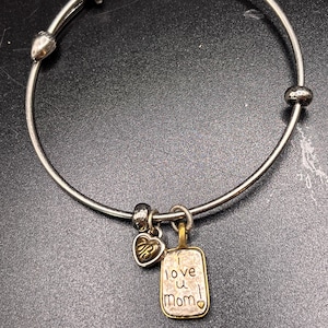 K&ouml;nnte beinhalten: Silberfarbenes Armband mit Herz- und rechteckigen Anh&auml;ngern. Der rechteckige Anh&auml;nger tr&auml;gt die Gravur "I love u Mom!". Das Armband hat einen Herzanh&auml;nger mit den Initialen "RB".