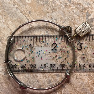 K&ouml;nnte beinhalten: Silberfarbenes Armreif-Armband mit Herzanh&auml;nger und einem rechteckigen Anh&auml;nger mit der Aufschrift "Born to Love, Home is where the heart is". Das Armband hat einen Durchmesser von ca. 7 cm.