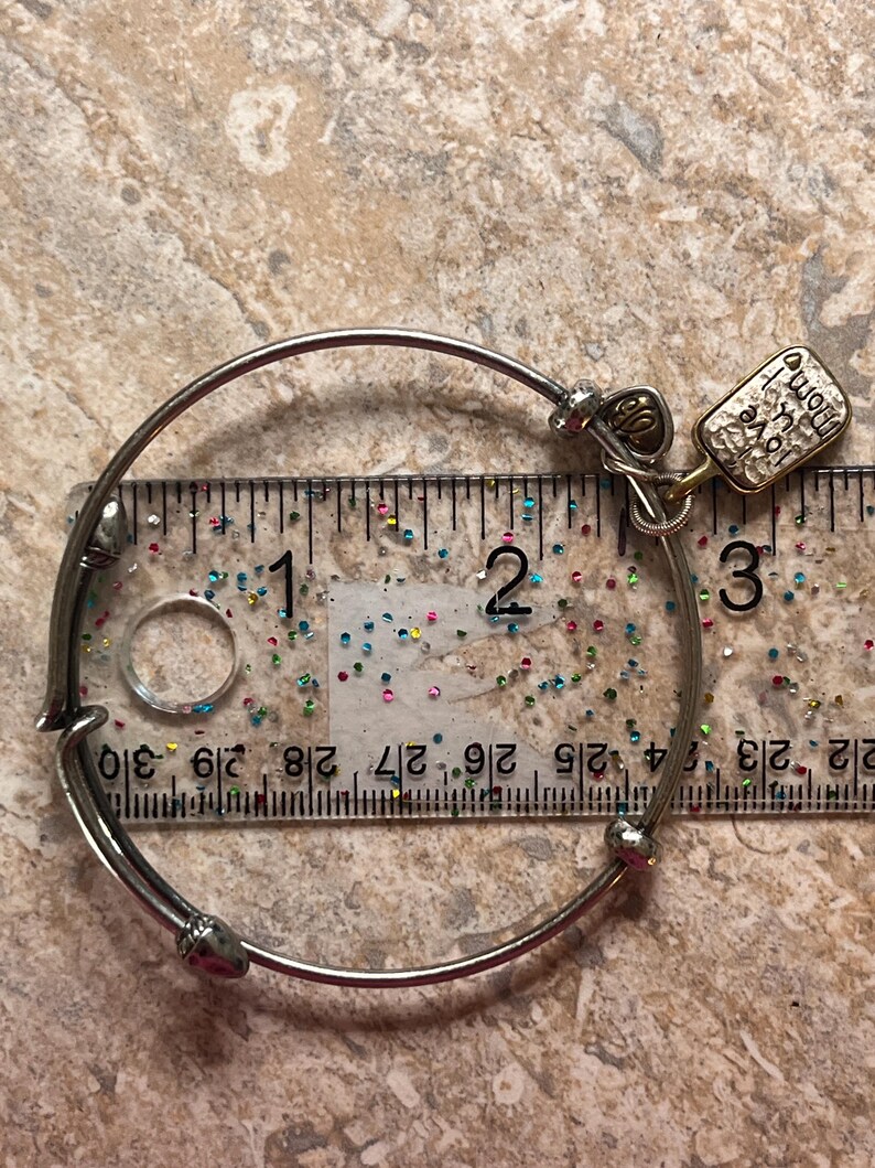 K&ouml;nnte beinhalten: Silberfarbenes Armreif-Armband mit Herzanh&auml;nger und einem rechteckigen Anh&auml;nger mit der Aufschrift "Born to Love, Home is where the heart is". Das Armband hat einen Durchmesser von ca. 7 cm.