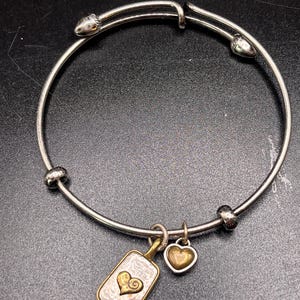 K&ouml;nnte beinhalten: Silberfarbenes Armreif-Armband mit zwei Anh&auml;ngern. Ein Anh&auml;nger ist ein rechteckiger Anh&auml;nger mit einem goldenen Herzdesign, der andere ein kleines goldenes Herz. Das Armband hat einen Schiebeverschluss.