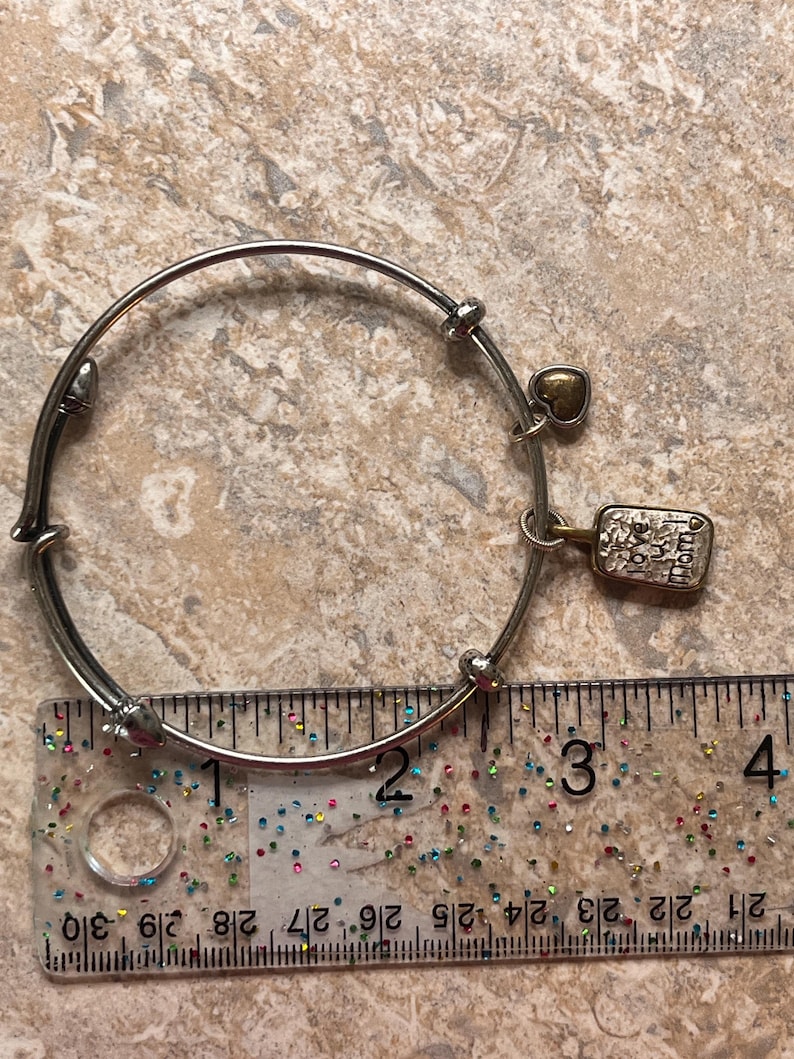 K&ouml;nnte beinhalten: Silberfarbenes Armreif-Armband mit einem kleinen goldenen Herzanh&auml;nger und einem rechteckigen Anh&auml;nger mit der Gravur "love you mom". Das Armband hat einen Durchmesser von ca. 7,6 cm.