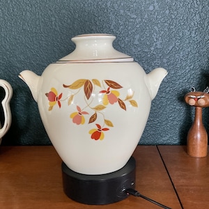 May include: A cream-colored ceramic vessel with two side handles and a lid, featuring a floral pattern in shades of yellow, orange, and brown. The vessel is positioned on a black circular rotating display stand with a power cord attached.