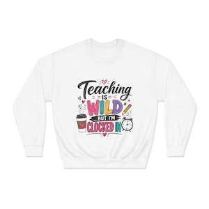 May include: A white long-sleeve sweatshirt featuring a colorful graphic print that reads Teaching is WILD but I'm CLOCKED IN. The design includes a coffee cup with a heart, a yellow pencil, and a black alarm clock surrounded by small pink hearts.