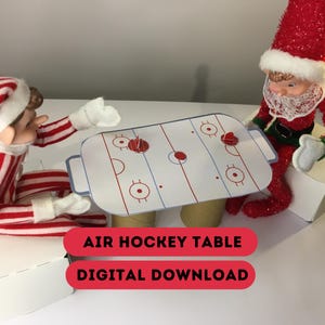 May include: A miniature air hockey table with red and blue markings, supported by cardboard tubes, is set up between two elf figurines. The table has red pucks and is labeled "AIR HOCKEY TABLE" and "DIGITAL DOWNLOAD" in red text.