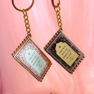 May include: Two rectangular keychains with gold chains. One keychain has a pink and floral design, the other has a black and floral design. Both keychains have the text "AND HE IS WITH YOU WHERE EVER YOU ARE".