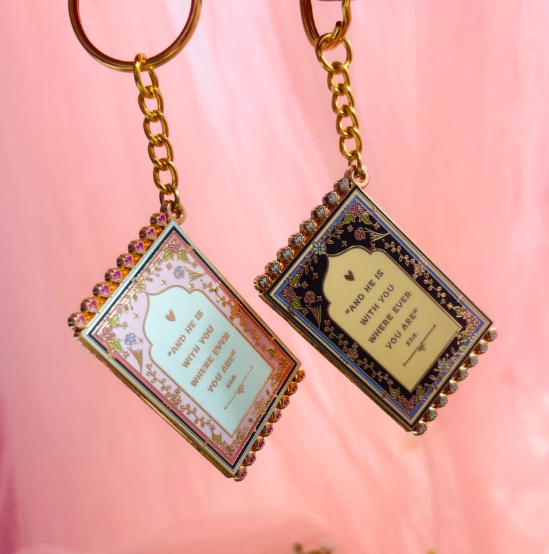 May include: Two rectangular keychains with gold chains. One keychain has a pink and floral design, the other has a black and floral design. Both keychains have the text "AND HE IS WITH YOU WHERE EVER YOU ARE".