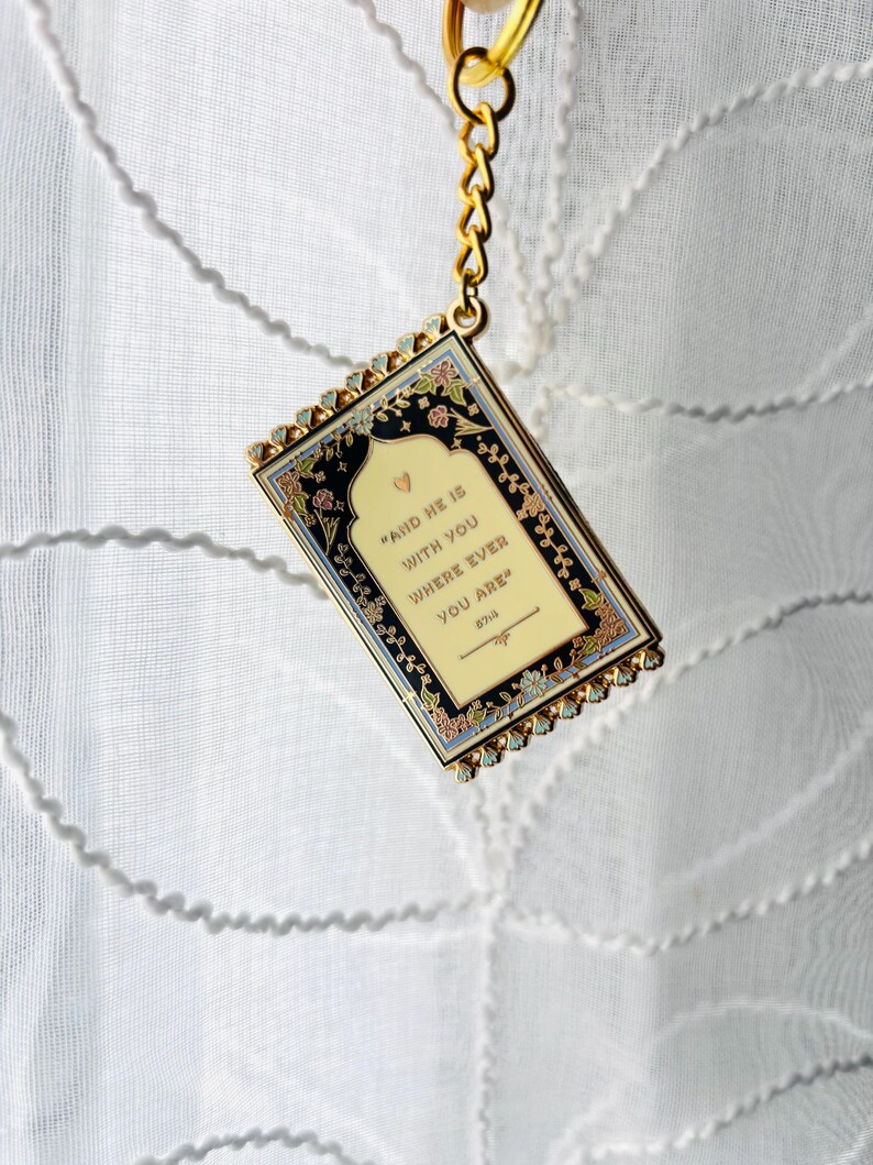 May include: A rectangular keychain with a gold chain and a decorative frame. The frame has a black background with floral accents and the text "AND HE IS WITH YOU WHERE EVER YOU ARE".