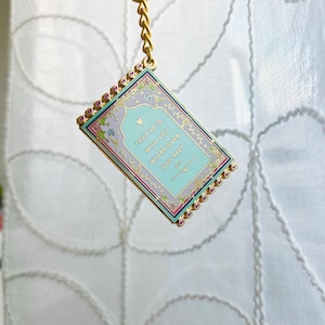May include: A rectangular keychain with a gold chain and clasp. The keychain features a light blue background with the text "AND HE IS WITH YOU WHERE EVER YOU ARE". The border is decorated with pink and gold accents.