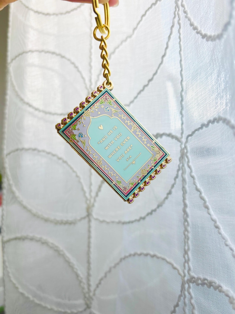 May include: A rectangular keychain with a gold chain and clasp. The keychain features a light blue background with the text "AND HE IS WITH YOU WHERE EVER YOU ARE". The border is decorated with pink and gold accents.
