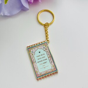 May include: Gold-colored keychain with a rectangular pendant. The pendant has a light blue background with the text "AND HE IS WITH YOU WHERE EVER YOU ARE". The edges are pink and gold, with floral accents.